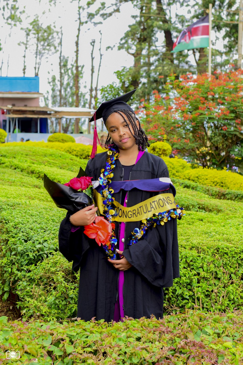 College graduate in Kenya standing outside holding flowers and she is wearing a purple cap and gown